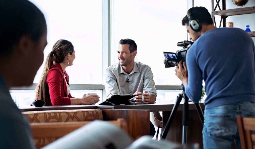 Happy businessman during corporate interview with female journalist. Manager answering question in office. Young woman at work as reporter with business man and cameraman shooting video for broadcast