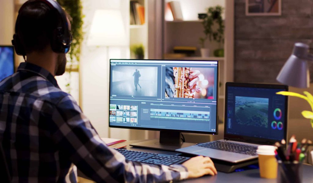 Film maker pointing at the monitor in home office while working on post production for a movie. Video editor wearing headphones.