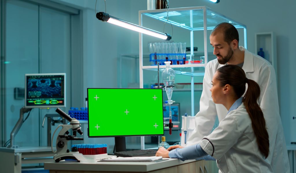Female research scientist has conversation with man bioengineer next to a desktop computer with green screen mock up. They look at chroma key pc template in a modern science laboratory.